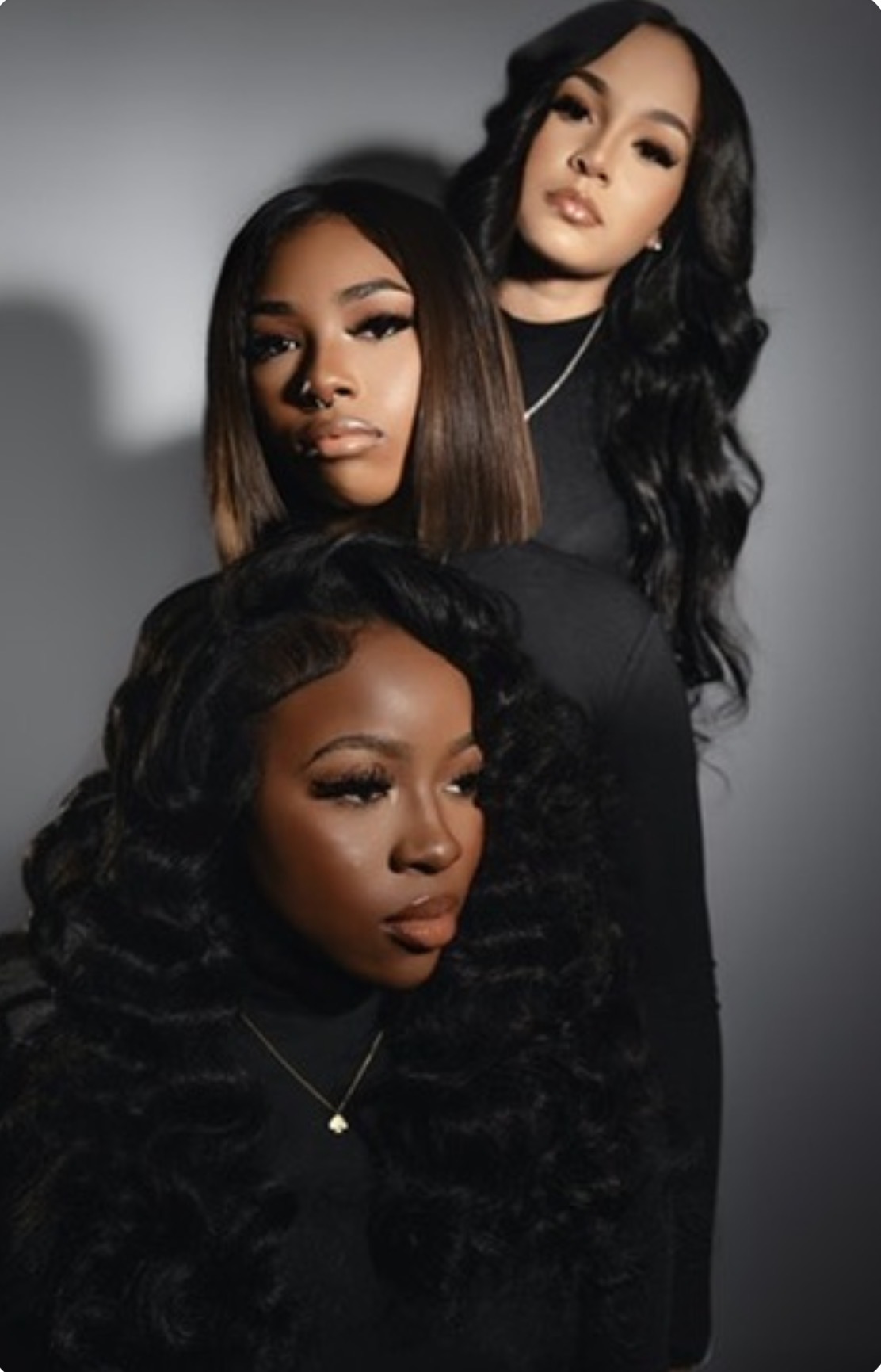Three women with long, dark hair posing together against a gray background, wearing black outfits.