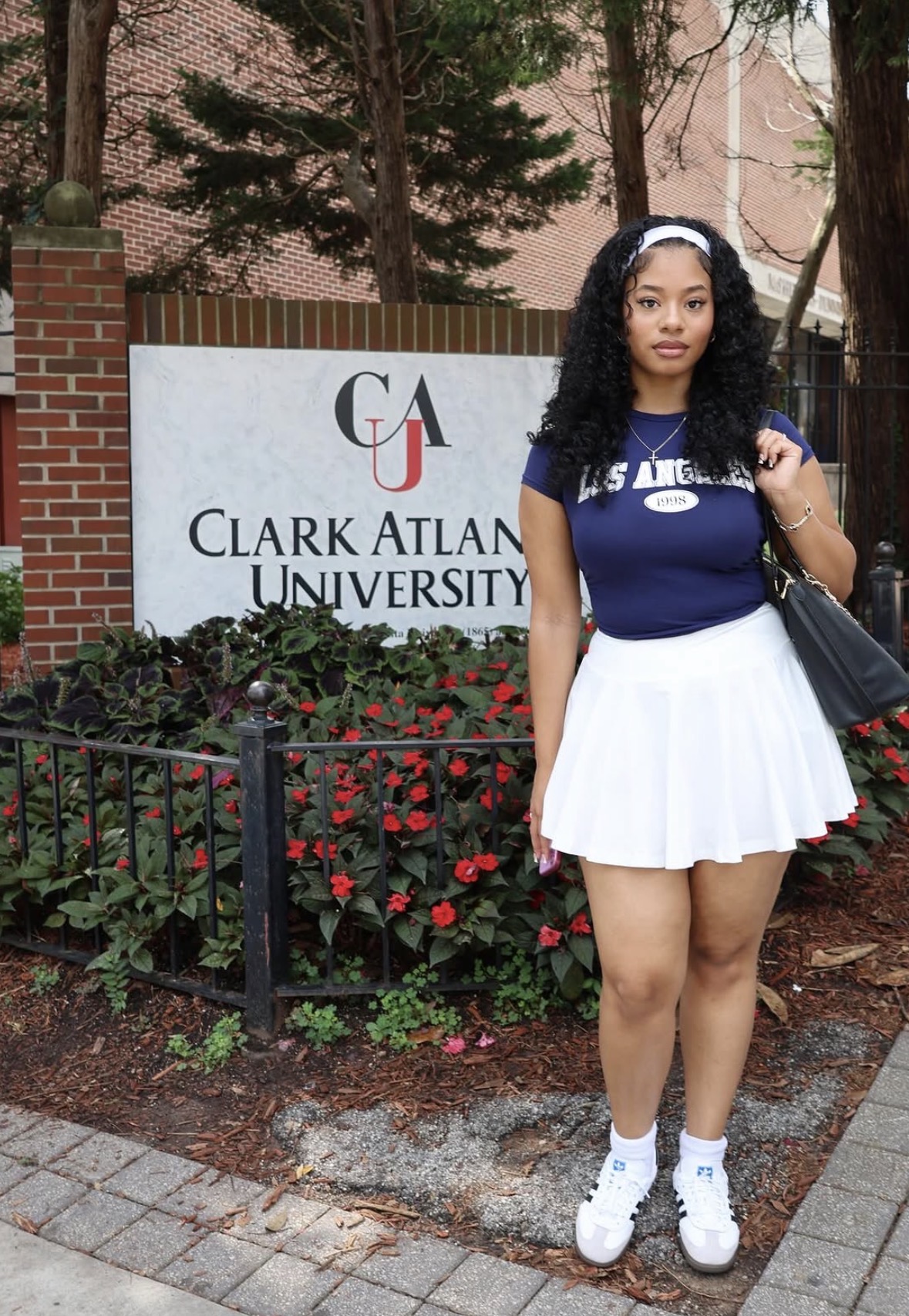 Young woman with curly hair in a blue shirt and white skirt standing outdoors near a university sign and flower bed.