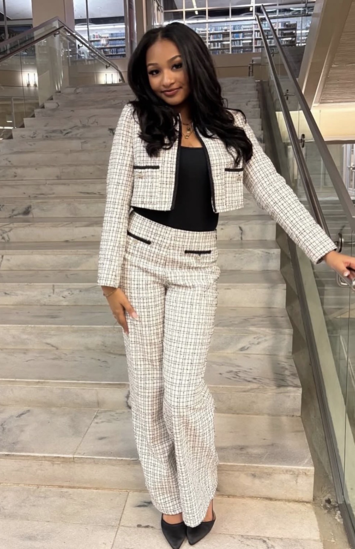 Woman in a checkered suit standing on stairs indoors, smiling, with glass railing and modern interior background.