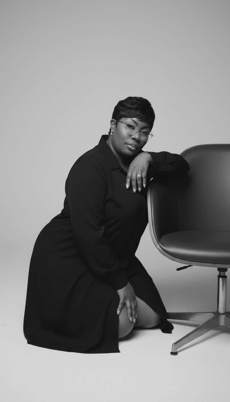 Person kneeling next to a chair, resting their arm on it, wearing glasses and a dark outfit.