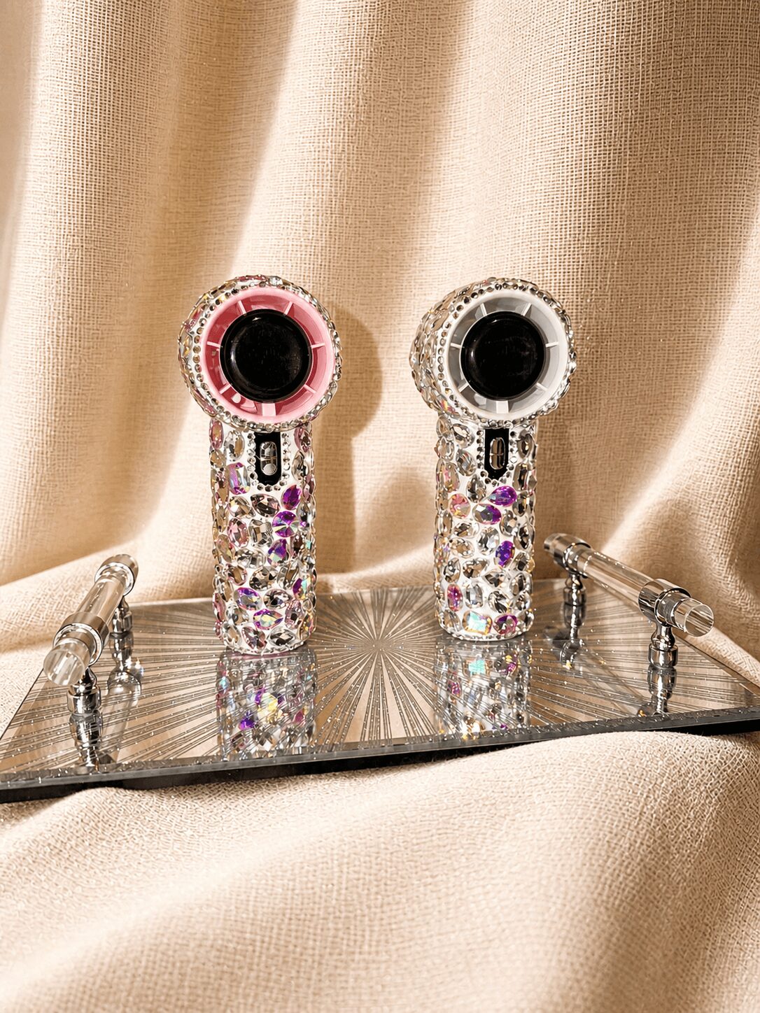 Two small, cylindrical devices with rhinestones and black centers on a reflective surface, against a beige fabric background.