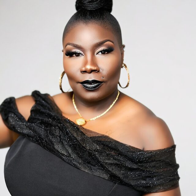Woman with dark lipstick, gold hoop earrings, and a black off-shoulder top, posing against a light background.