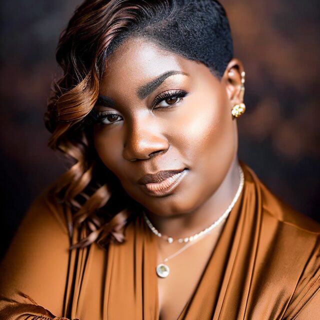 Close-up portrait of a woman with styled hair, wearing earrings, a necklace, and a brown satin top, looking at the camera.