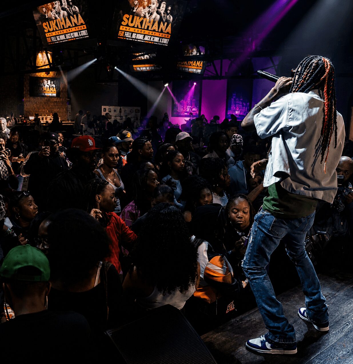 Performer with long braids and white jacket on stage, audience watching in a dark, colorful concert venue.