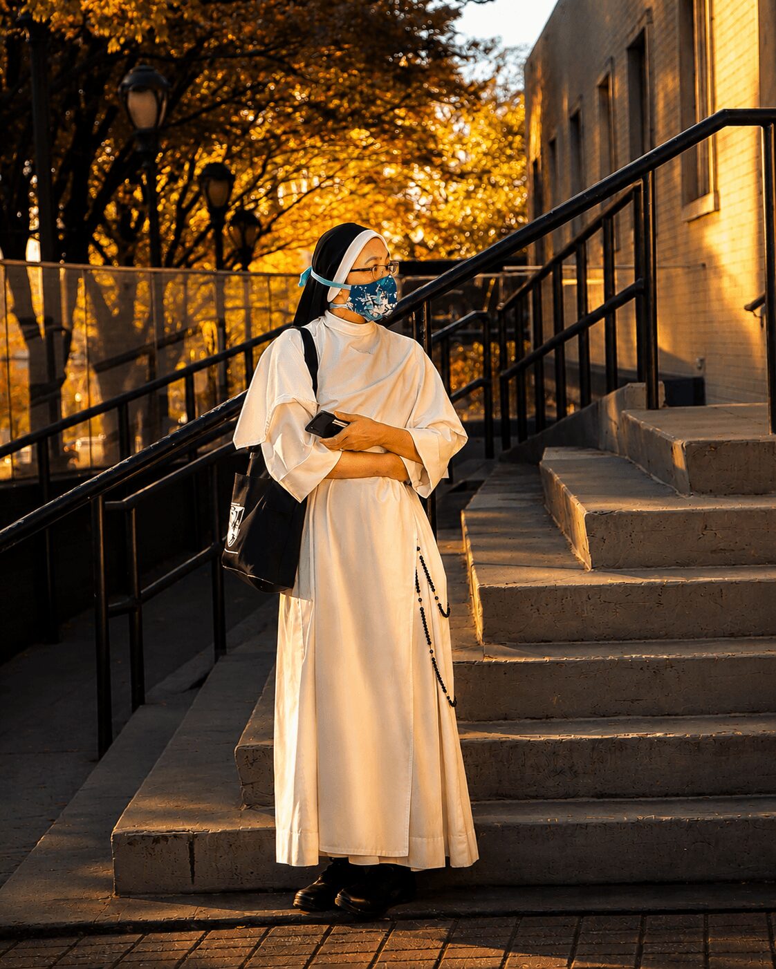 Person in a long white dress and face mask standing on stairs outside during sunset, with trees and building in background.