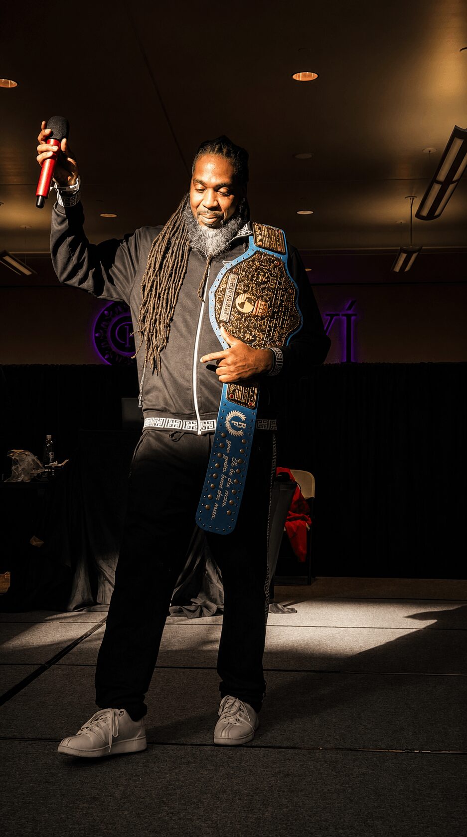 Man with dreadlocks holding a trophy and a sash, standing on stage with a dark background.