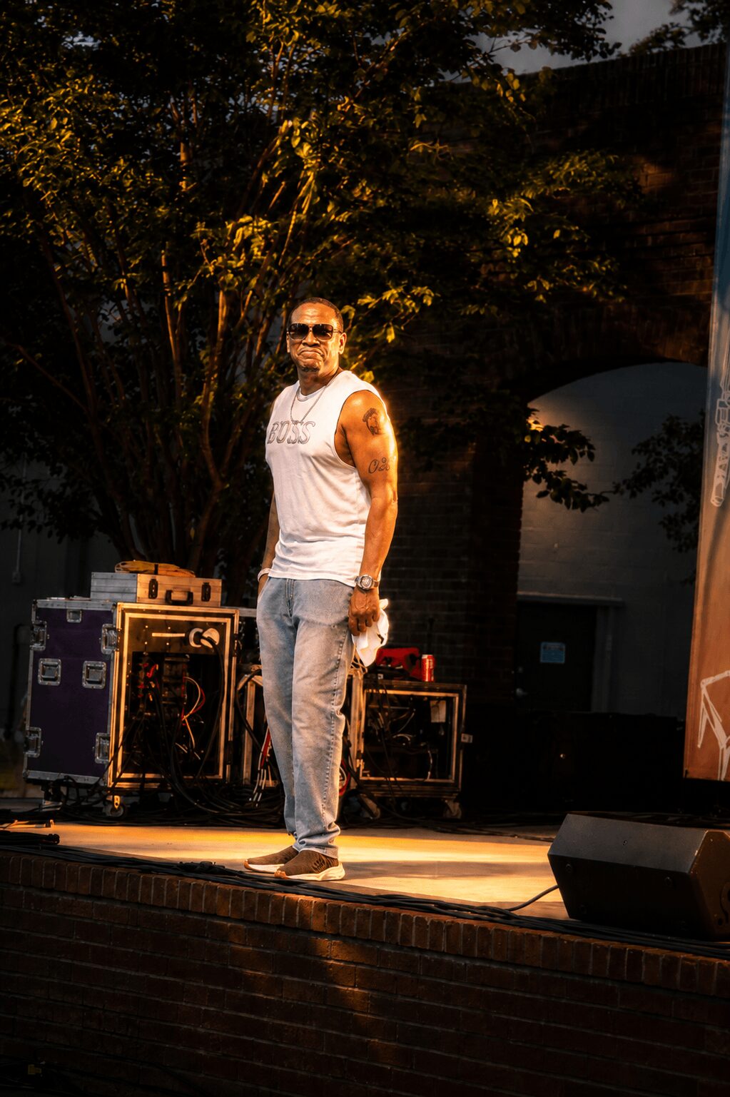 Man standing on stage outdoors, wearing sunglasses, sleeveless shirt, and jeans, with trees and equipment behind him.