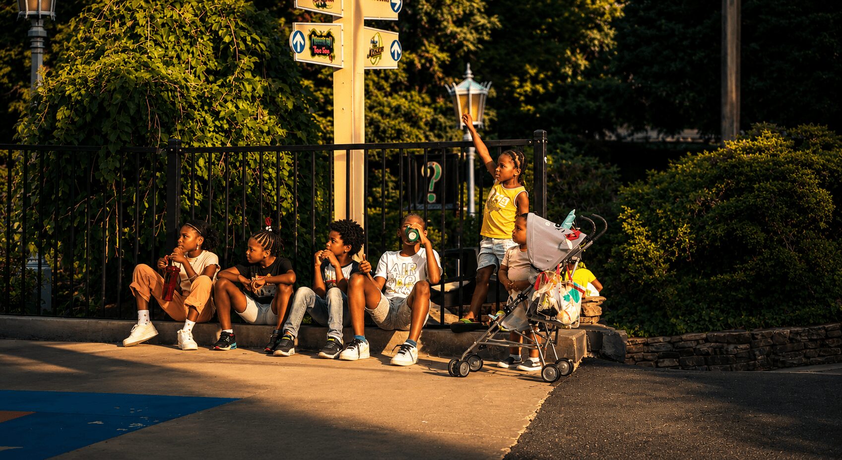 Group of children sitting and standing near a fence with trees in the background.