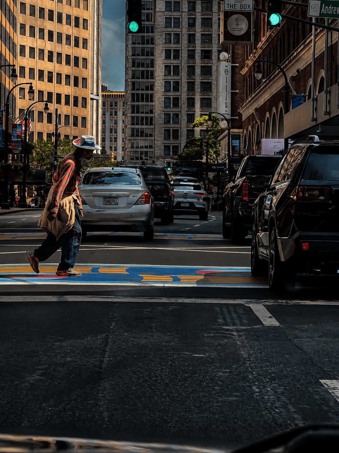 Person crossing street with buildings and cars in city background, traffic lights overhead, sunny day.