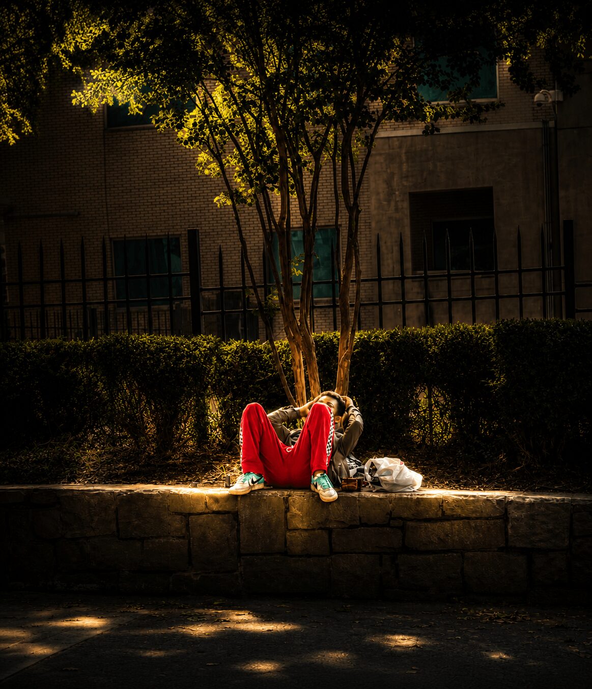 Person sitting on a low wall under a tree at night, illuminated by a spotlight, with a bag nearby.