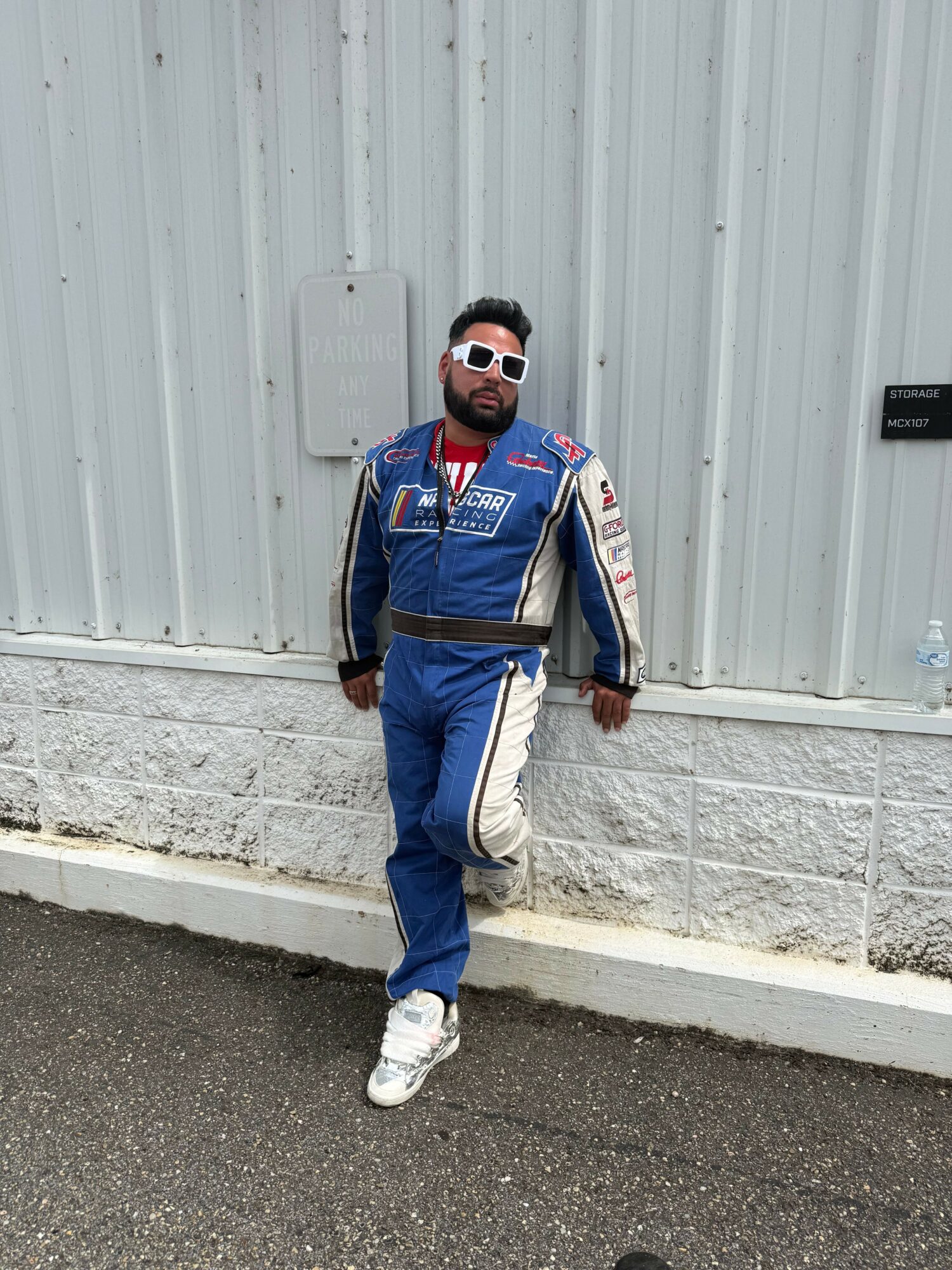 Person standing outdoors against a metal wall, wearing sunglasses, a racing suit, and sneakers, with hands at sides.