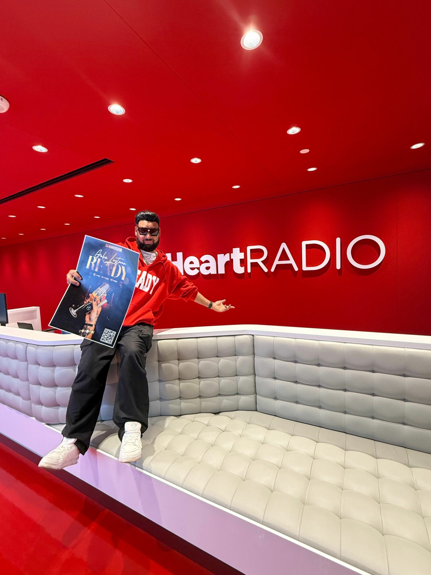 Person sitting on a white couch holding a magazine, with a red wall and 'iHeartRadio' logo behind.