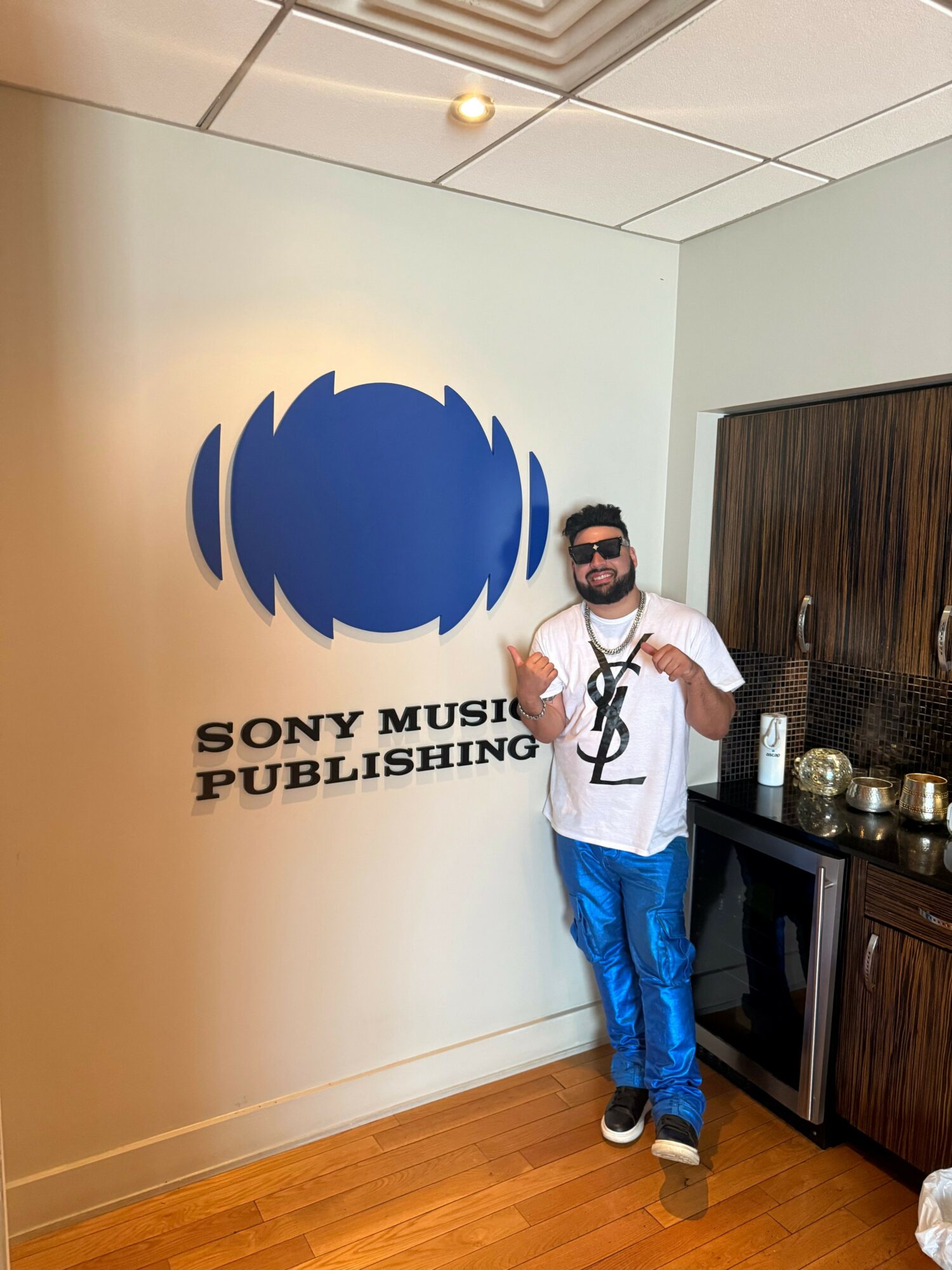 Man standing next to a wall with a Sony Music Publishing logo, holding a phone, in a room with wooden and tiled floors.