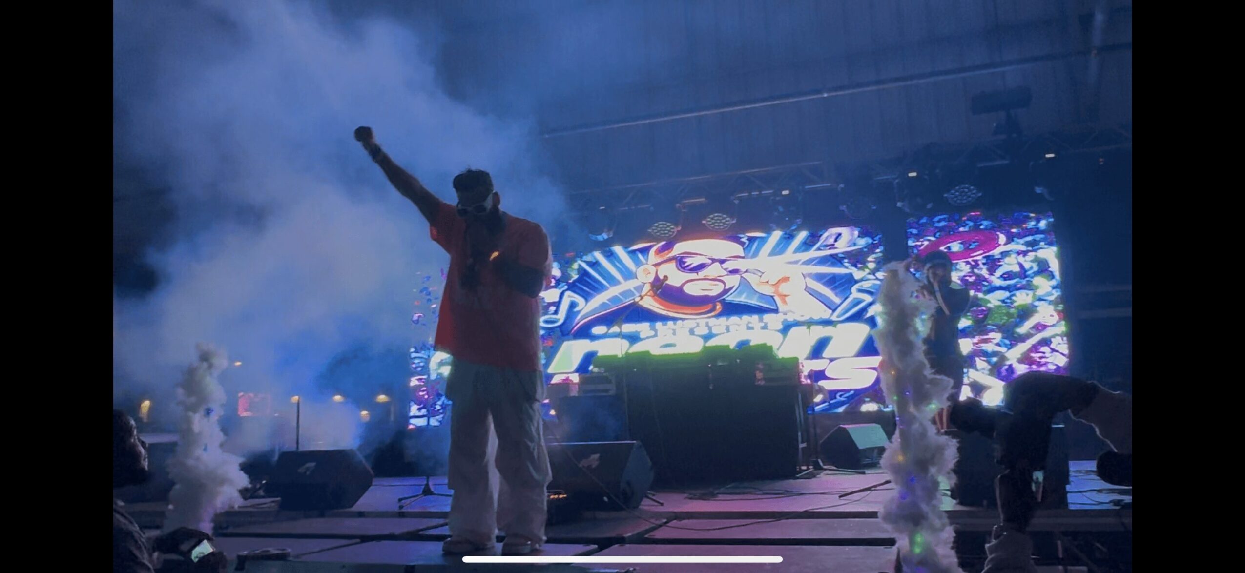 Performer on stage with raised arm, colorful background, smoke effects, and other performers present.
