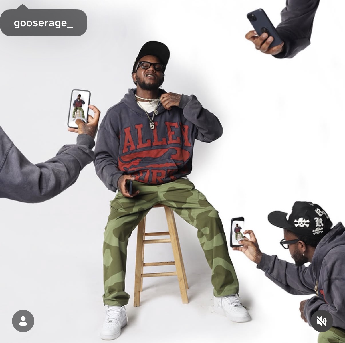 Man sitting on a stool, surrounded by three people taking photos with smartphones, wearing a hoodie and camouflage pants.