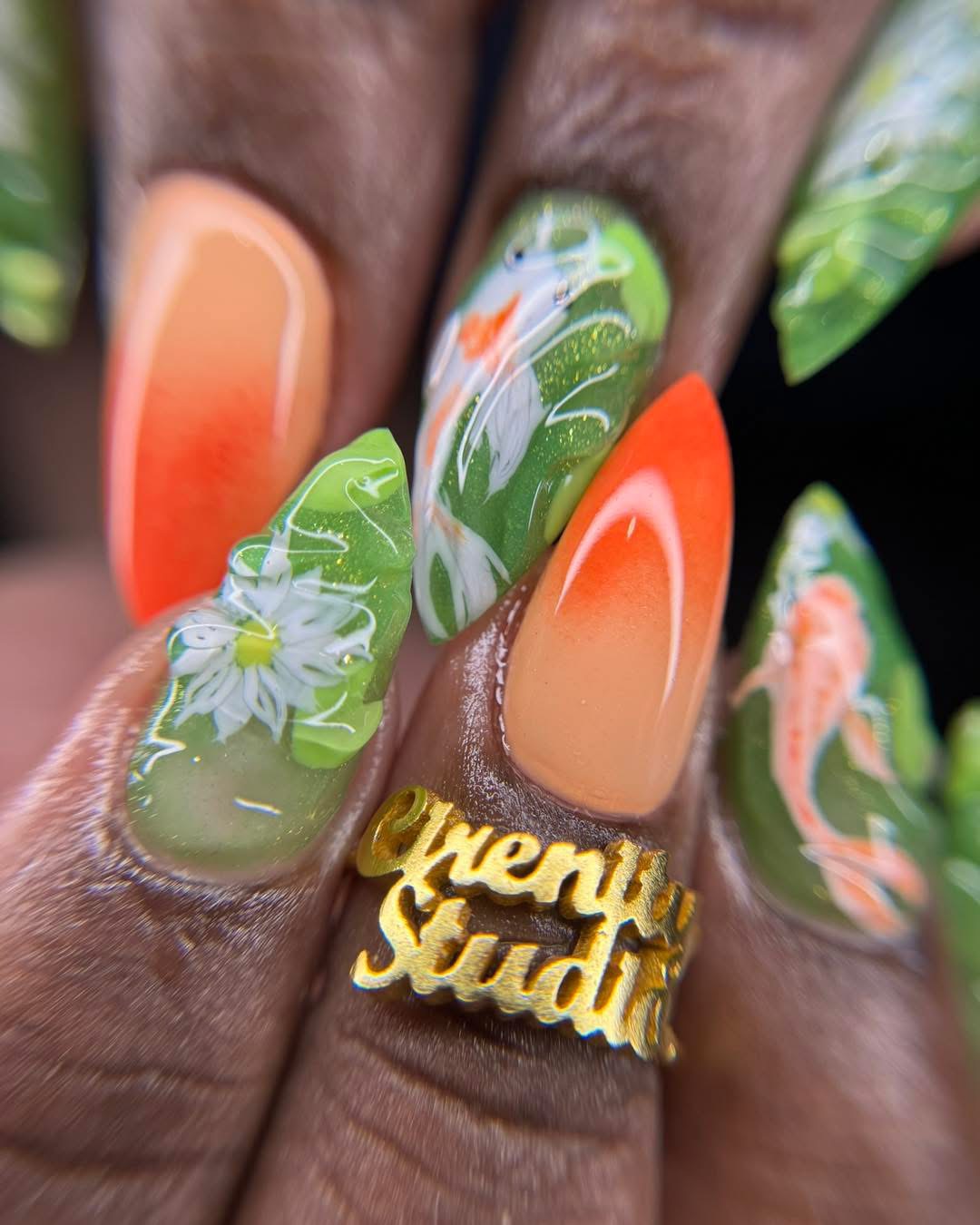 Nail art with floral and abstract designs in green, orange, and white, and a gold ring with text.