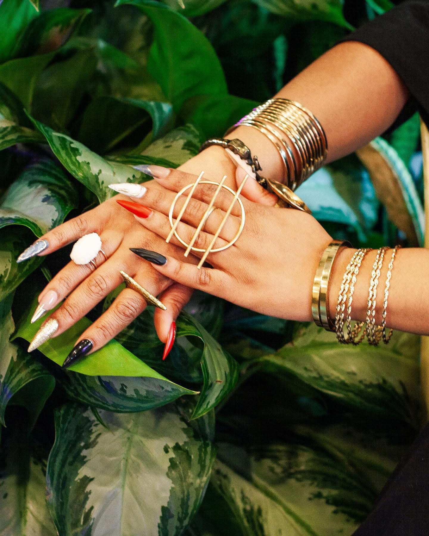 Close-up of hands with jewelry and manicured nails resting on green leaves.