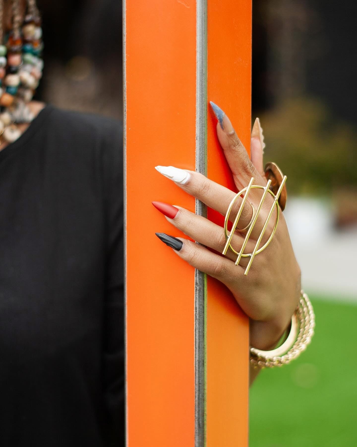 Hand with decorated nails and jewelry holding an orange pole outdoors, background blurred.