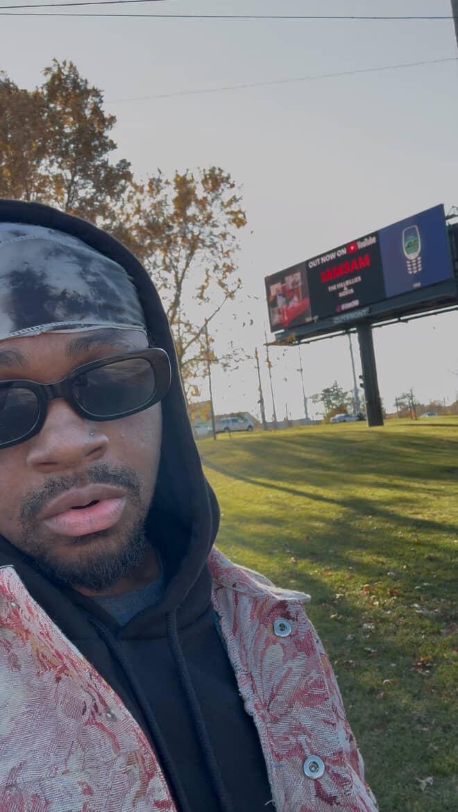 Part of a person's face wearing sunglasses and a hoodie, outdoors with trees and a billboard in the background.