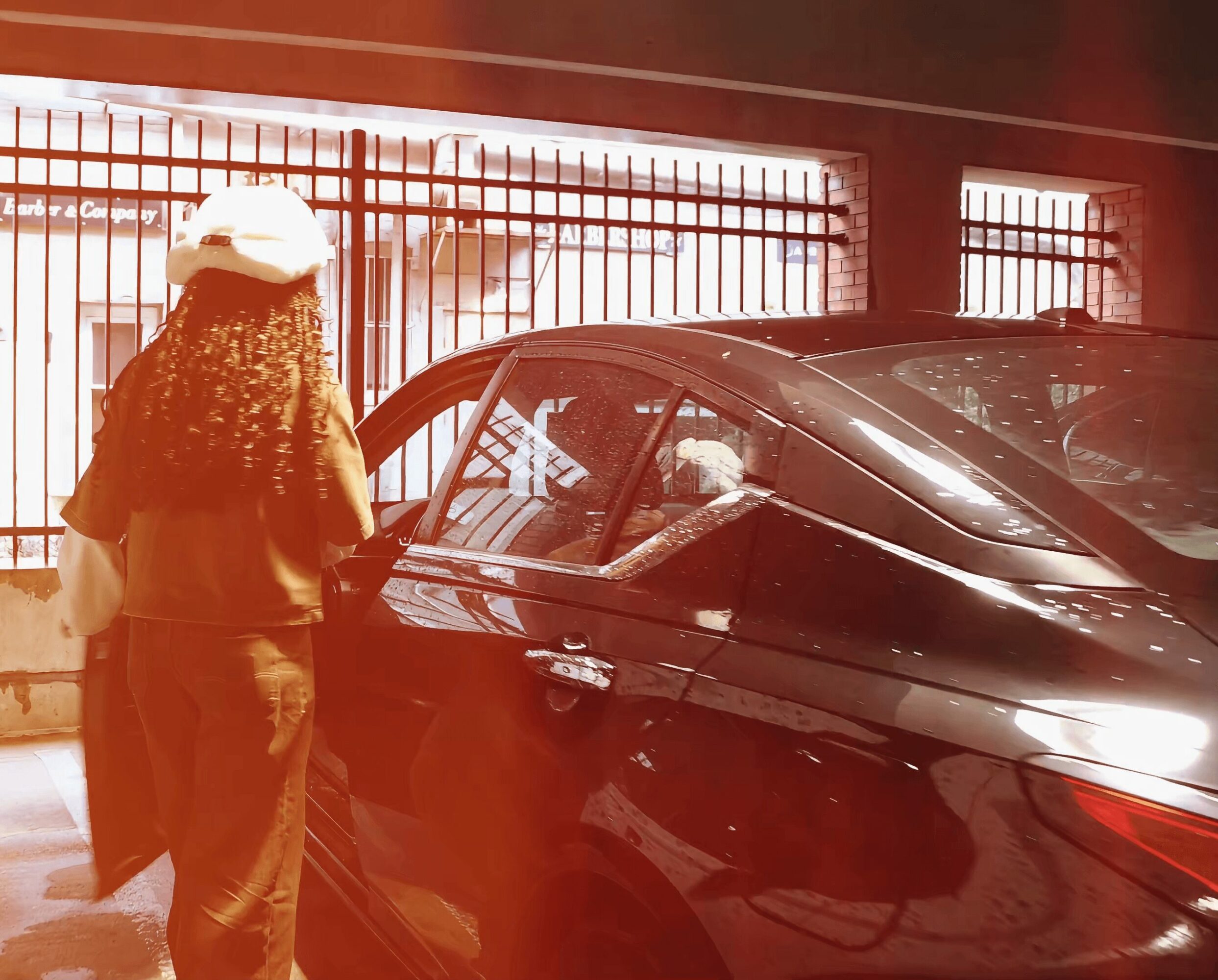Person with curly hair standing outside a black car, near a fence with vertical bars, in a parking area.