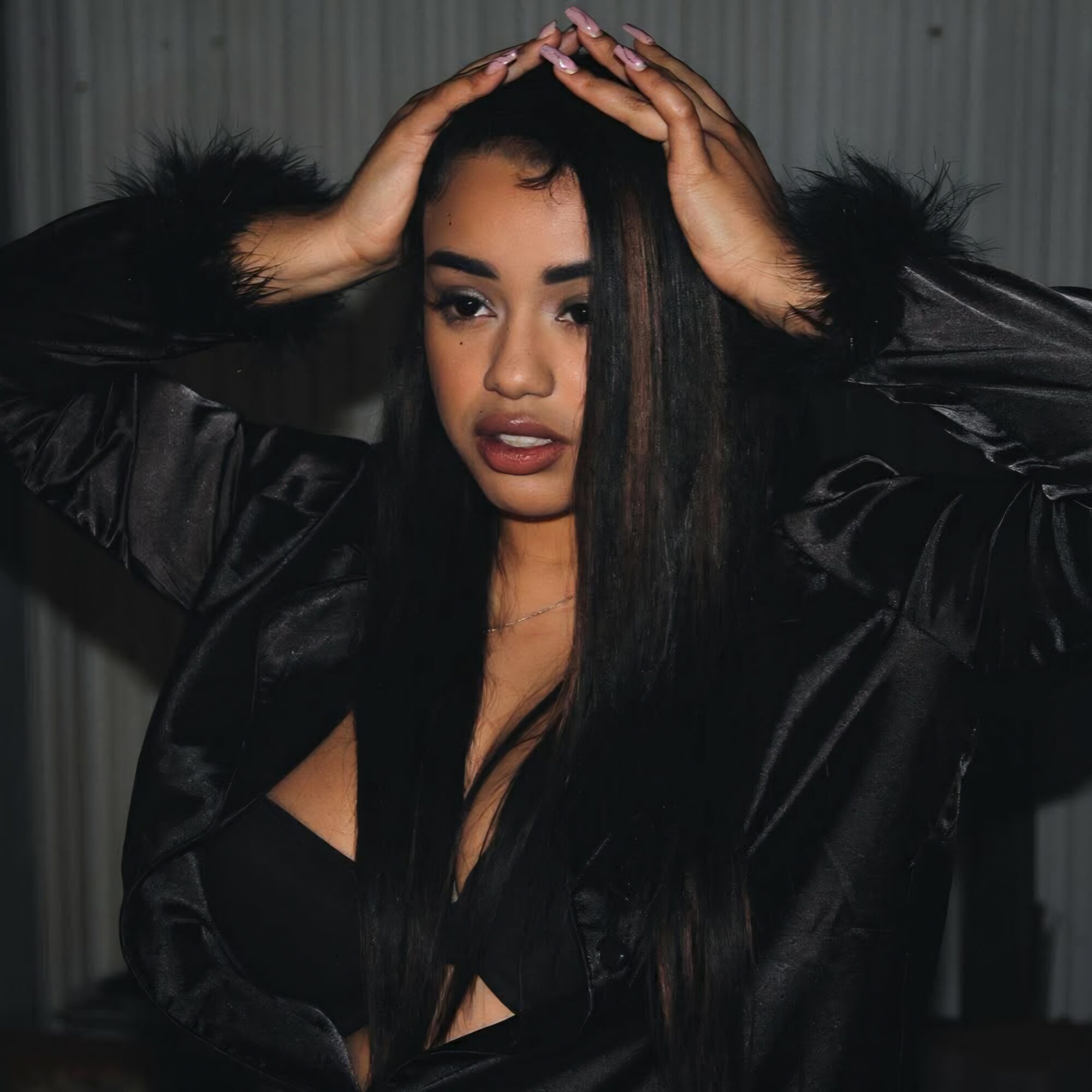 Woman with long black hair, wearing a black jacket, holding her head with both hands, looking at camera.