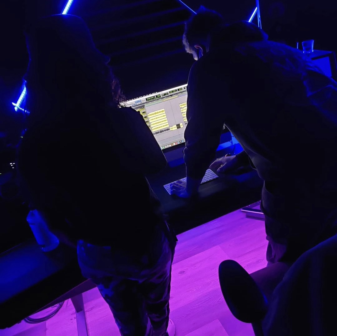 Two people are working on a computer in a dark room with blue lighting, leaning over a desk.
