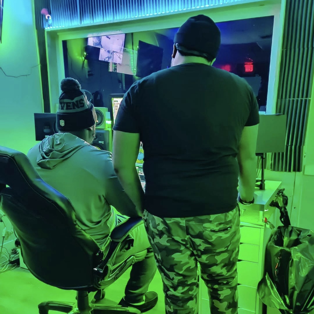 Two people in a room with green lighting, one sitting at a computer and the other standing nearby, facing away.