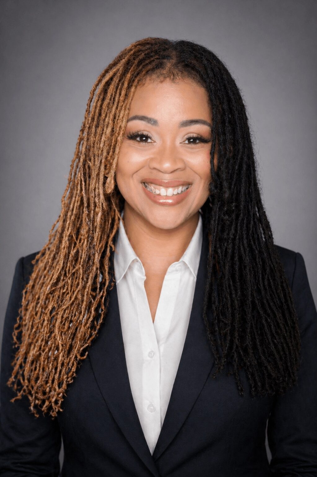 Portrait of a smiling woman with long, styled hair wearing a dark blazer and white shirt against a gray background.