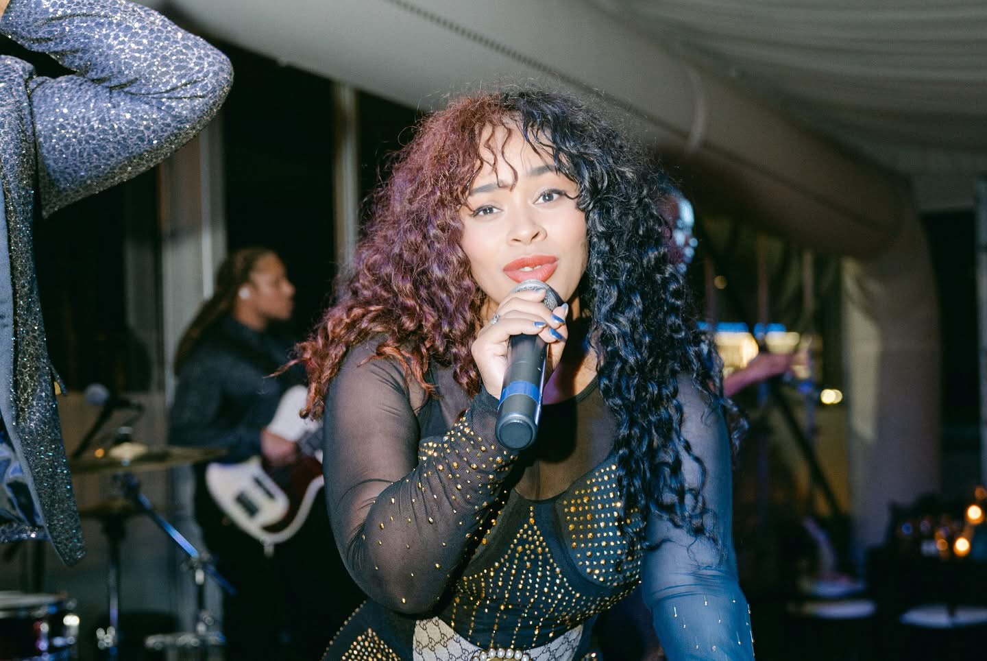 Female singer with curly hair holding a microphone performs on stage, with band members in the background.