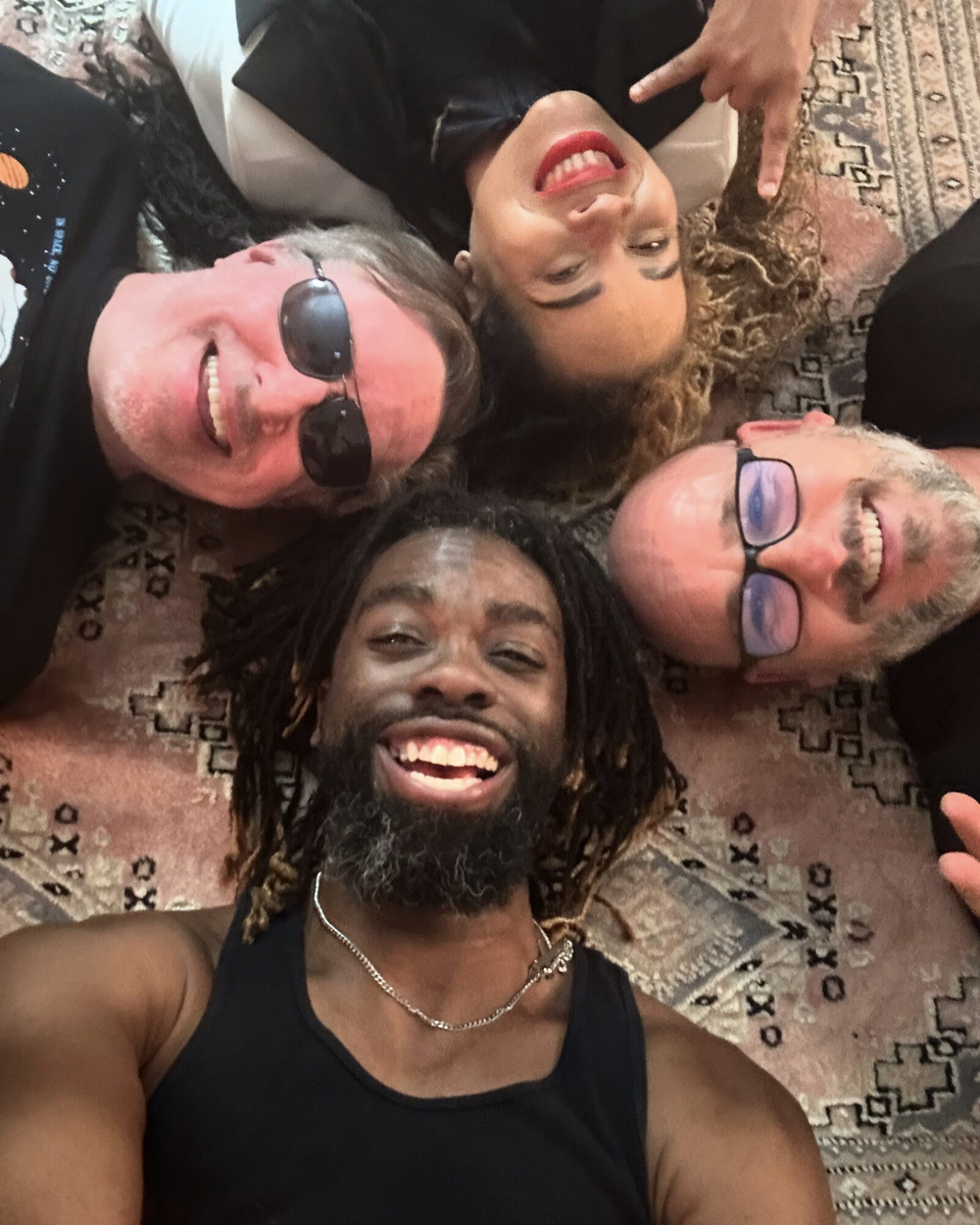 Four people lying on a patterned rug, smiling and making peace signs, with one person wearing sunglasses and another with glasses.