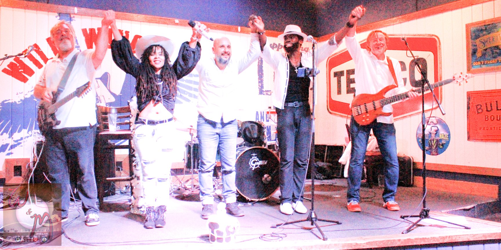 Group of six people performing on stage with musical instruments and microphones, some raising hands, colorful background, stage setup.