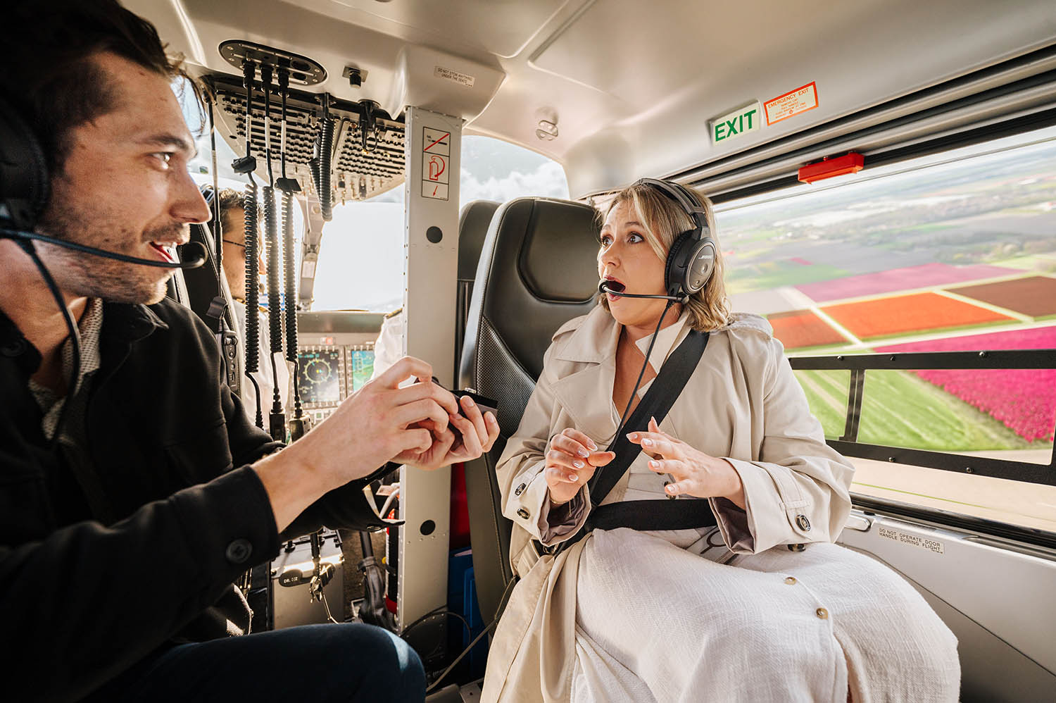 Two people sitting inside a helicopter, talking and wearing headsets, with a colorful landscape visible through the window.