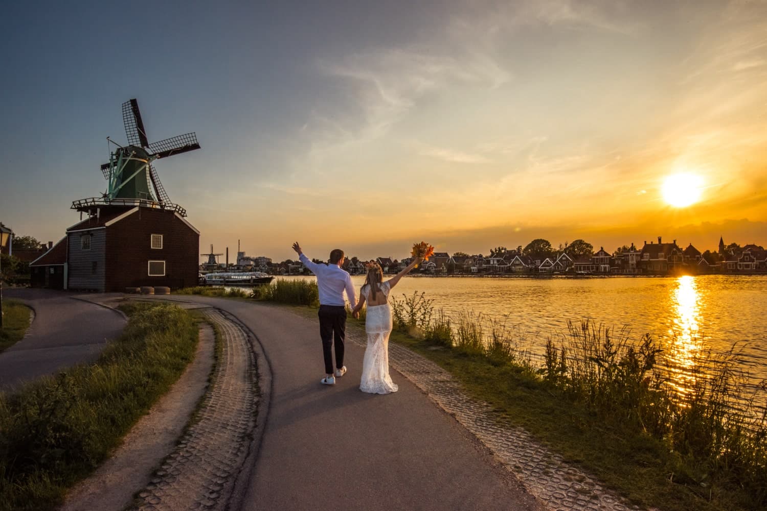 A couple holding hands walks along a river at sunset, with a windmill on the left and a city skyline in the distance.