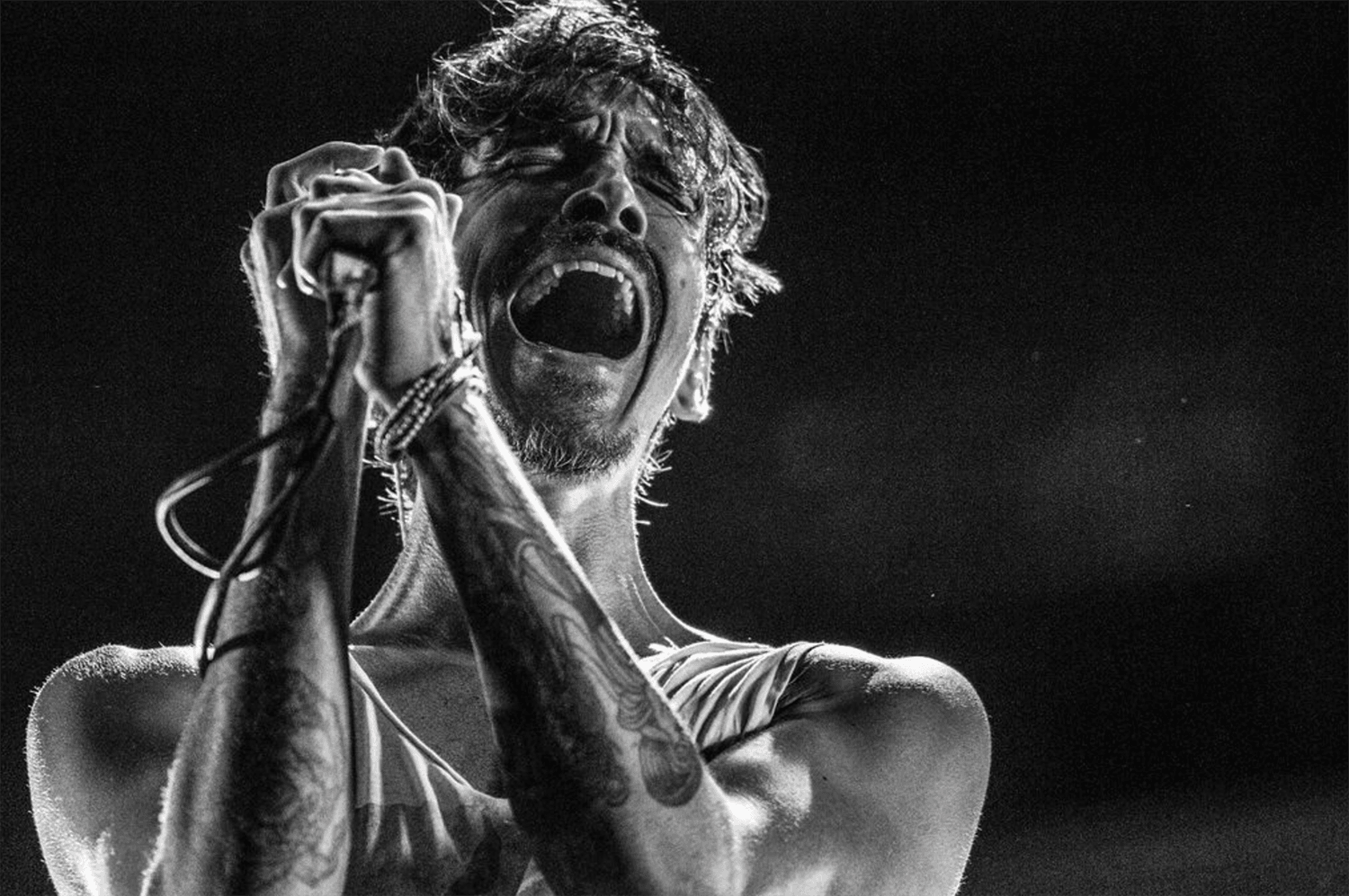 Person singing passionately into a microphone, with curly hair and tattoos, in black and white.