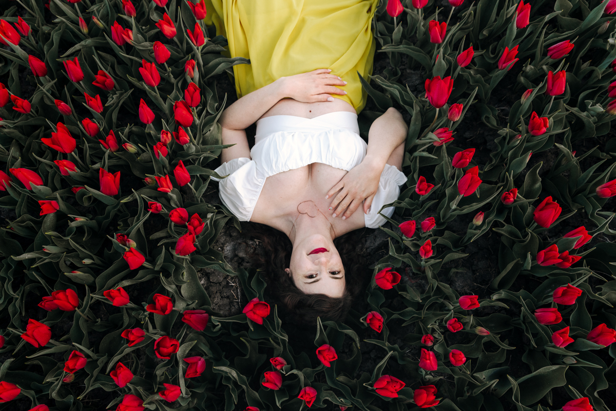 Woman lying among red tulips, wearing a yellow dress and white top, with her eyes closed and hands on her stomach.