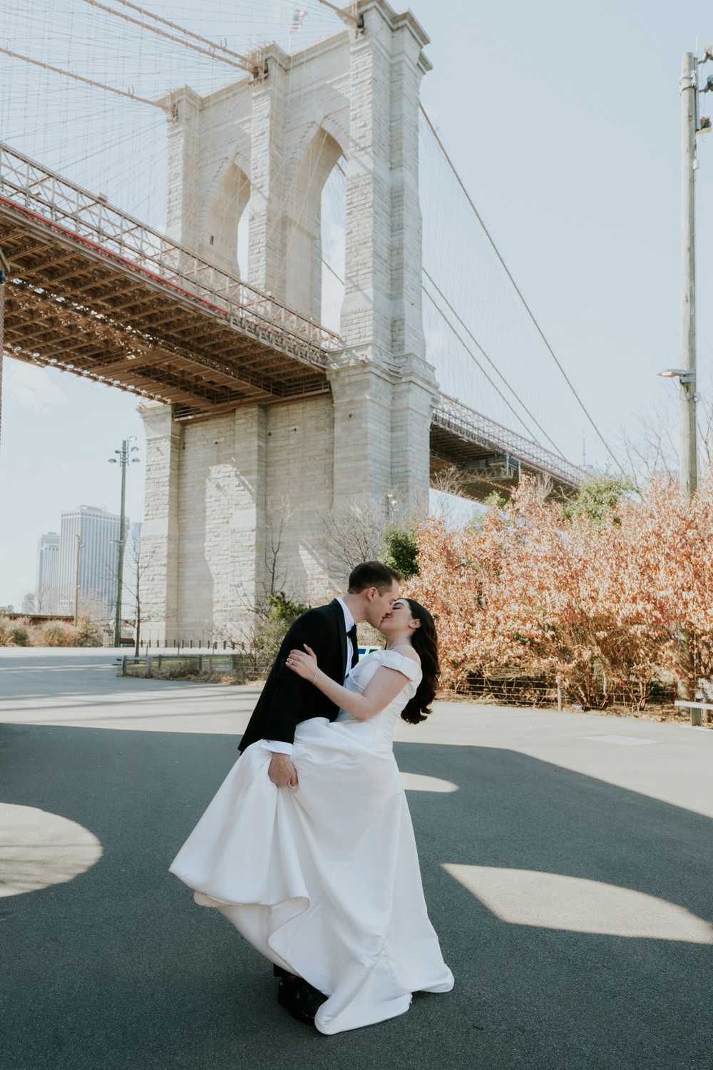 Couple dancing under a bridge with a cityscape in the background, trees with autumn leaves, and clear sky.