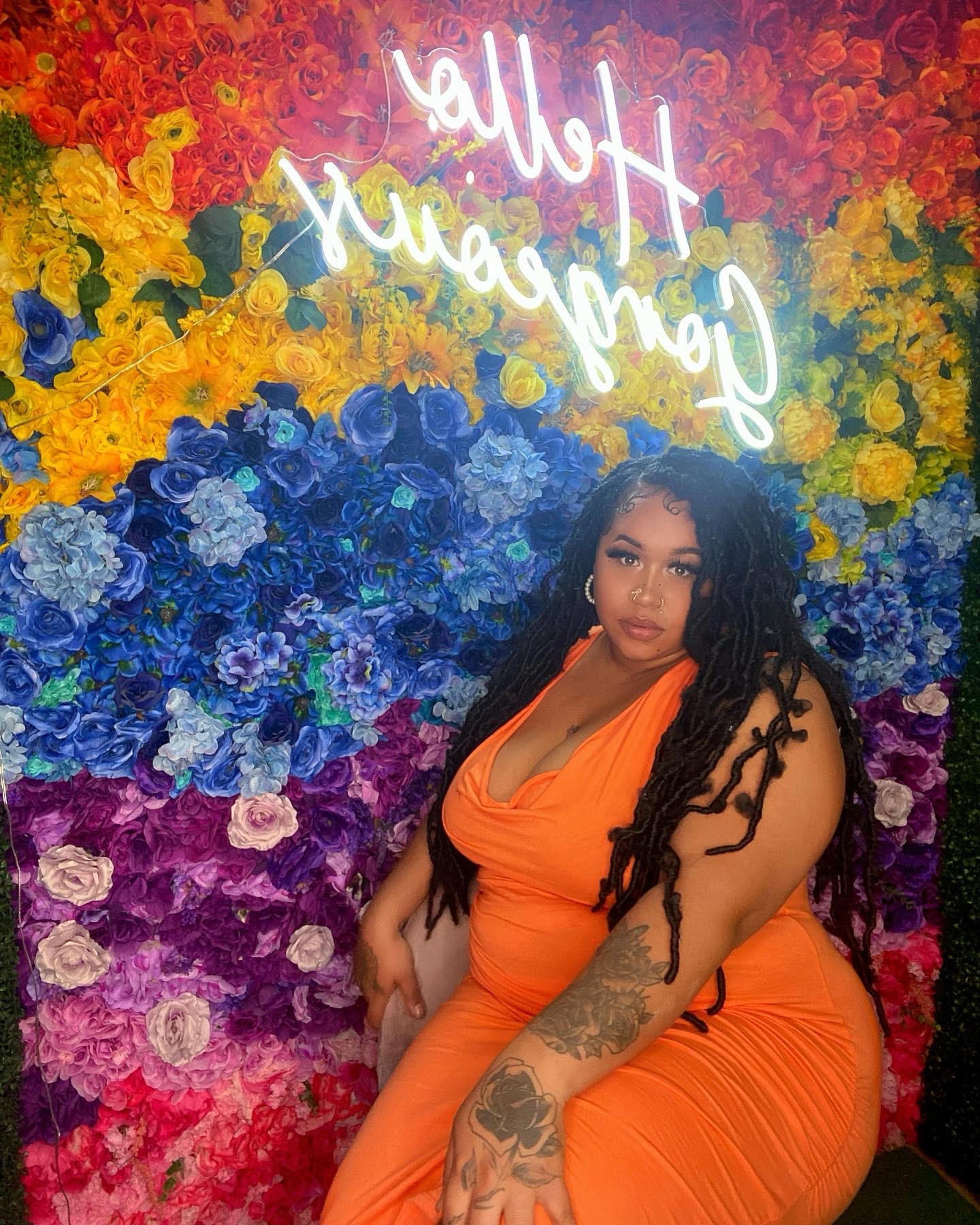 Woman with long curly hair in an orange dress, sitting in front of a colorful flower wall with a neon sign.