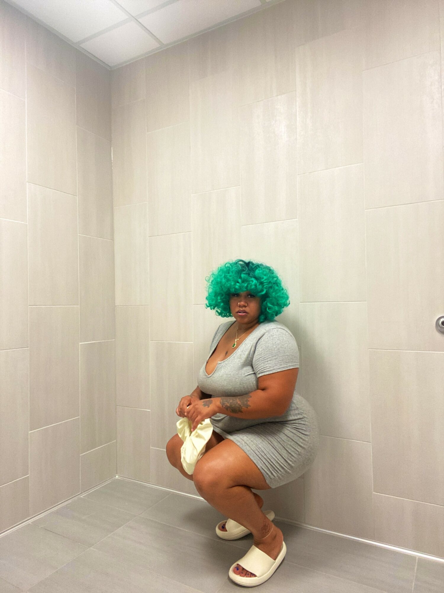 Person with bright green curly hair squatting in a beige tiled room, wearing a gray dress and white shoes.
