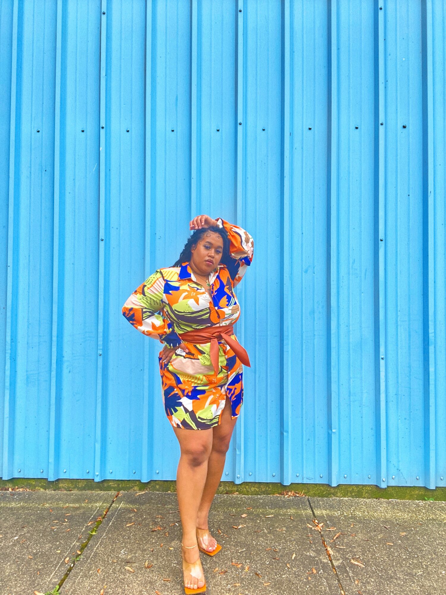 Woman in colorful dress standing against a blue corrugated metal wall, with one hand on her head and the other on her waist.