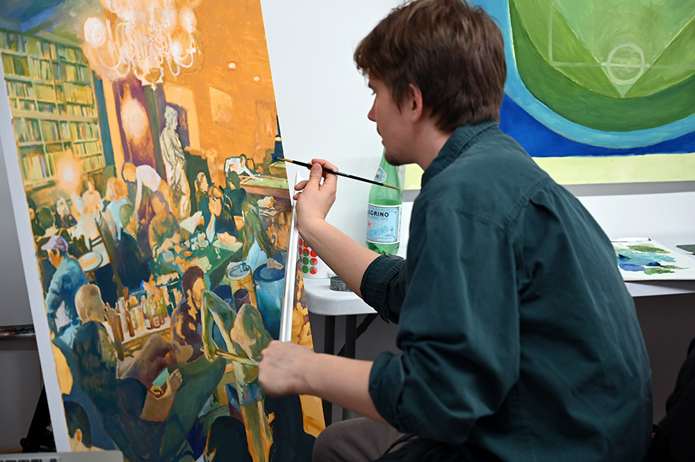 Young man painting a detailed scene of a lively indoor gathering with people, a chandelier, and warm lighting.