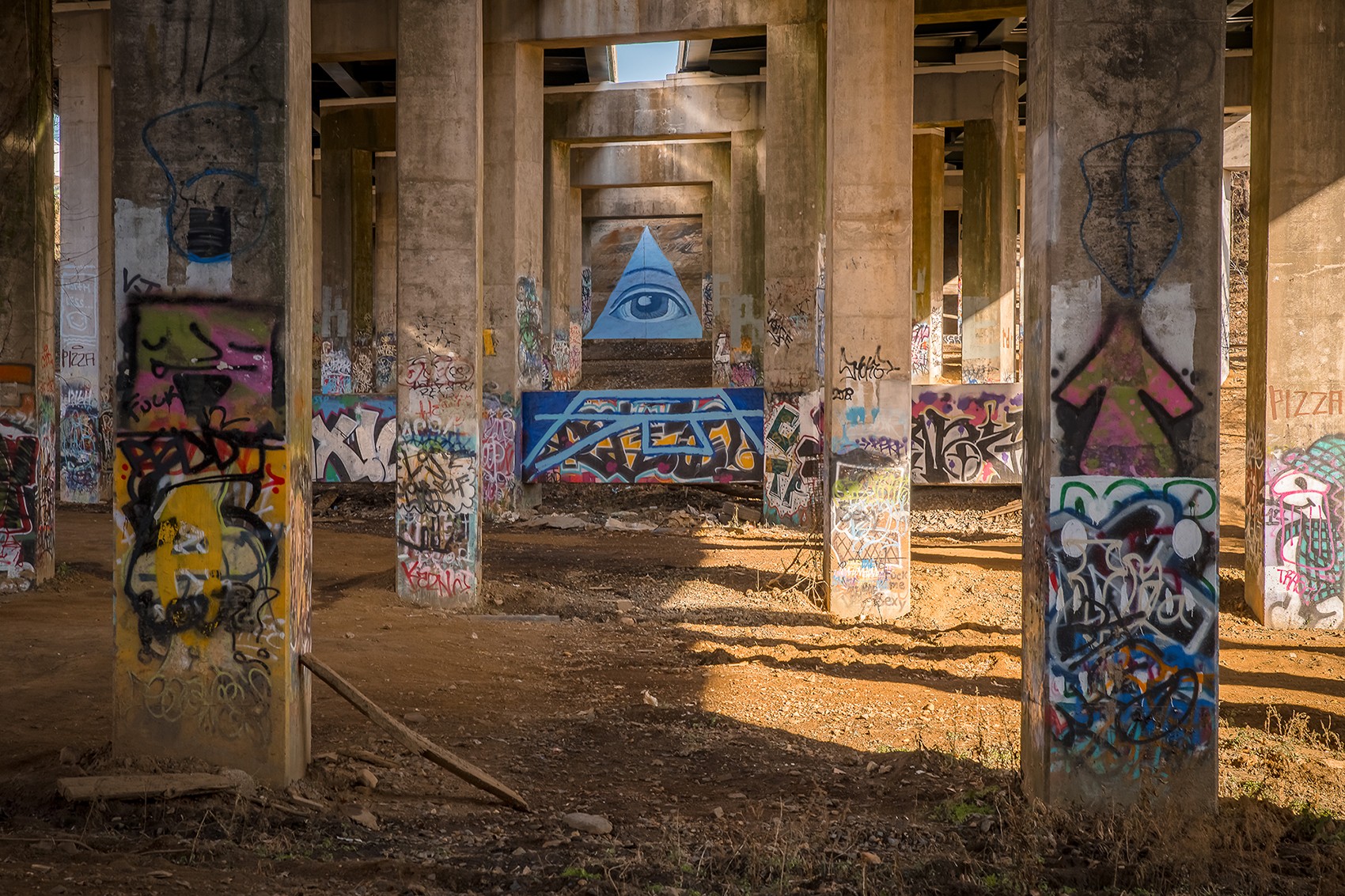 Under a bridge with concrete pillars covered in colorful graffiti, sunlight filters through gaps, illuminating the ground.