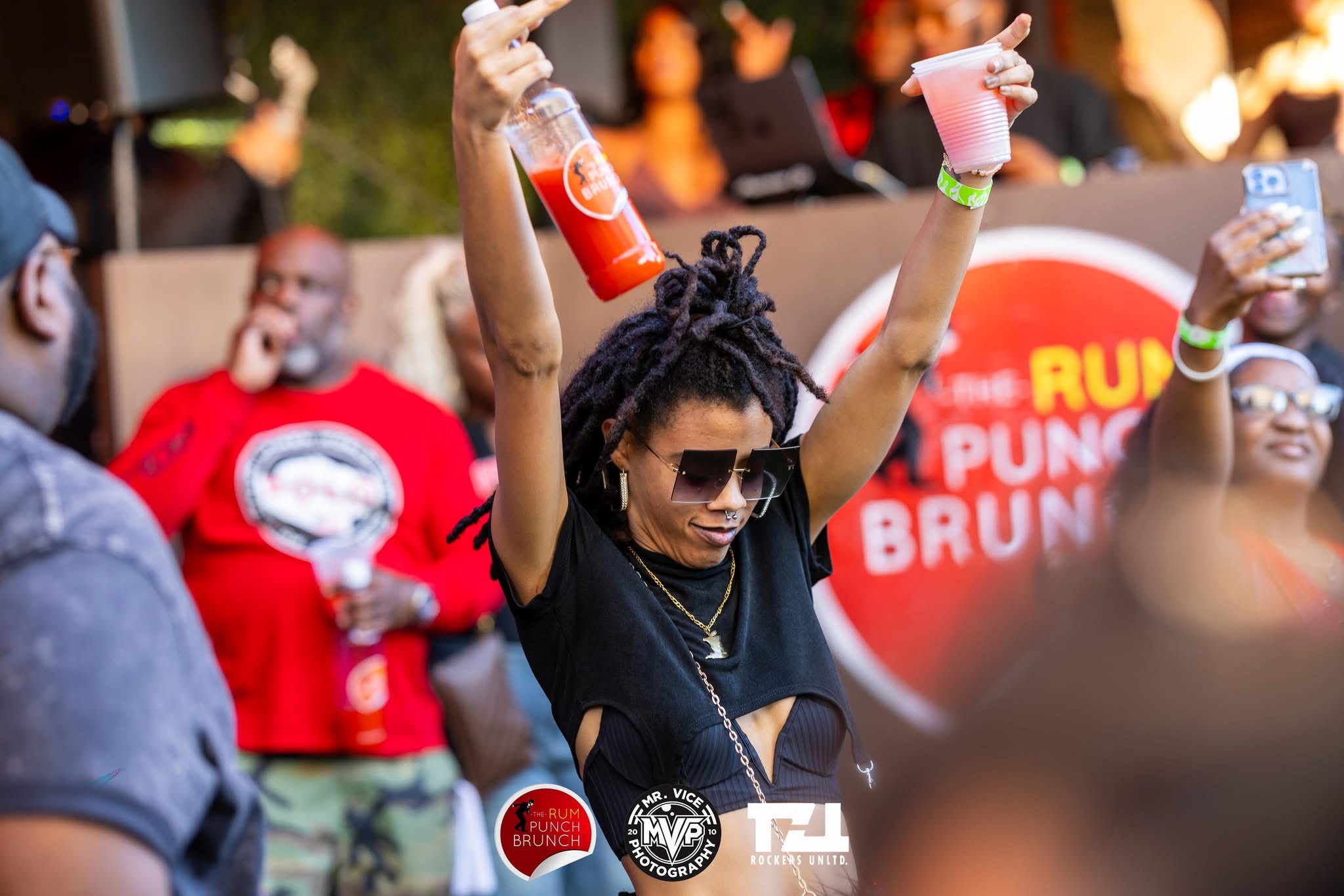 Person with dreadlocks and sunglasses raising two drinks at an outdoor event, with a crowd and a banner in the background.
