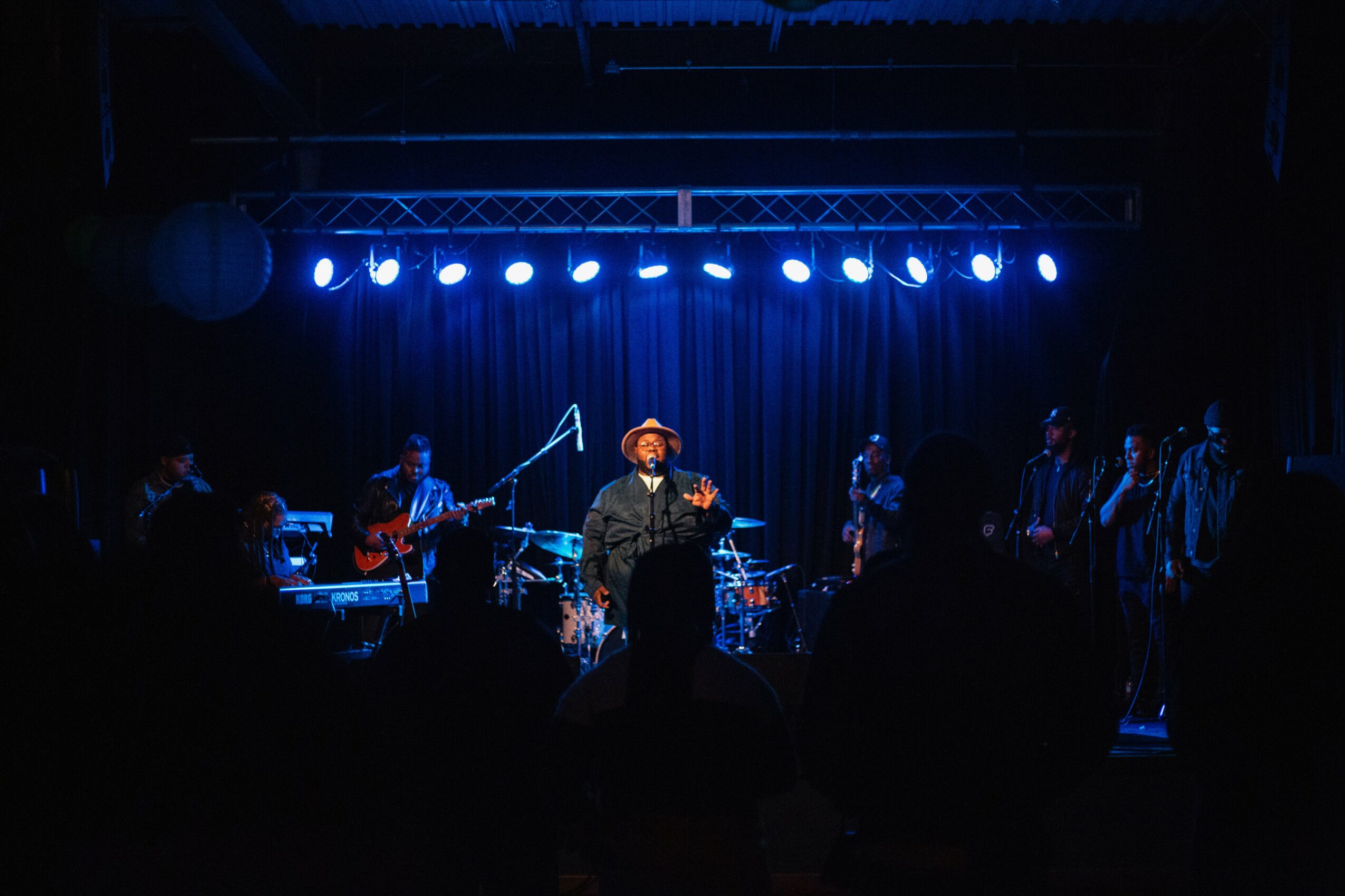 Stage with musicians performing under blue lights, audience silhouettes in foreground, dark background.