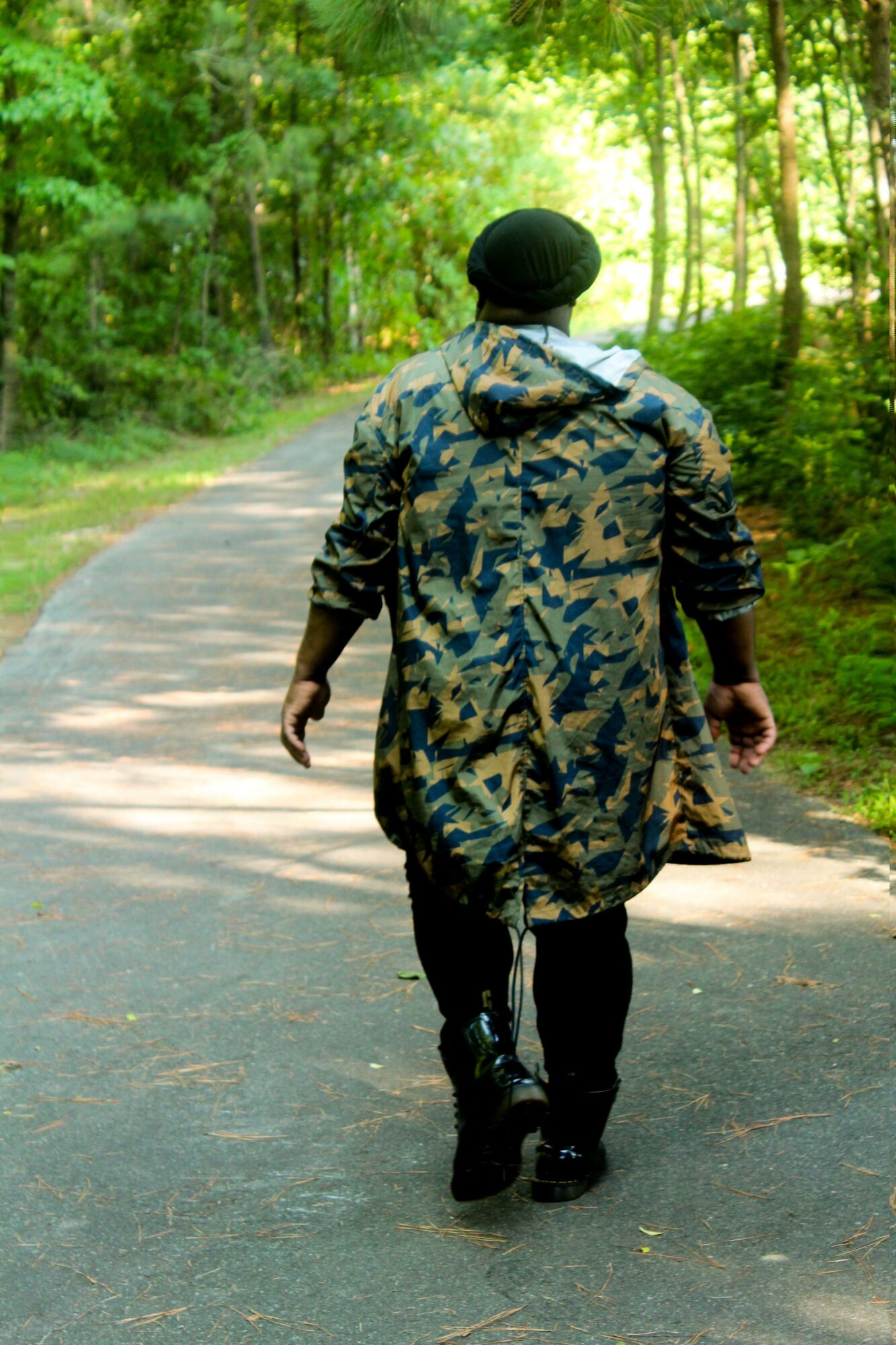 Person in camouflage coat walking on a forest path with trees and sunlight, back view.