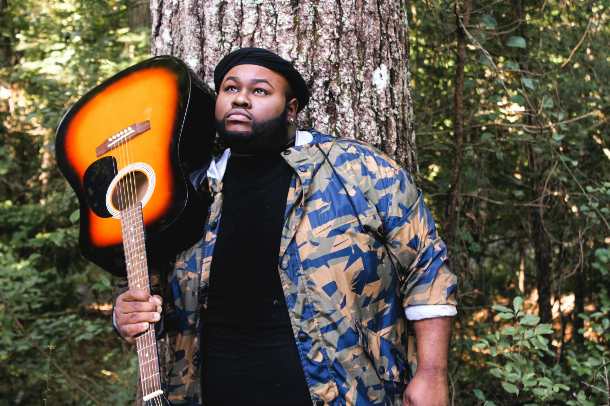 Person standing outdoors in a forest holding an acoustic guitar on their shoulder, wearing a camouflage jacket and black beanie.