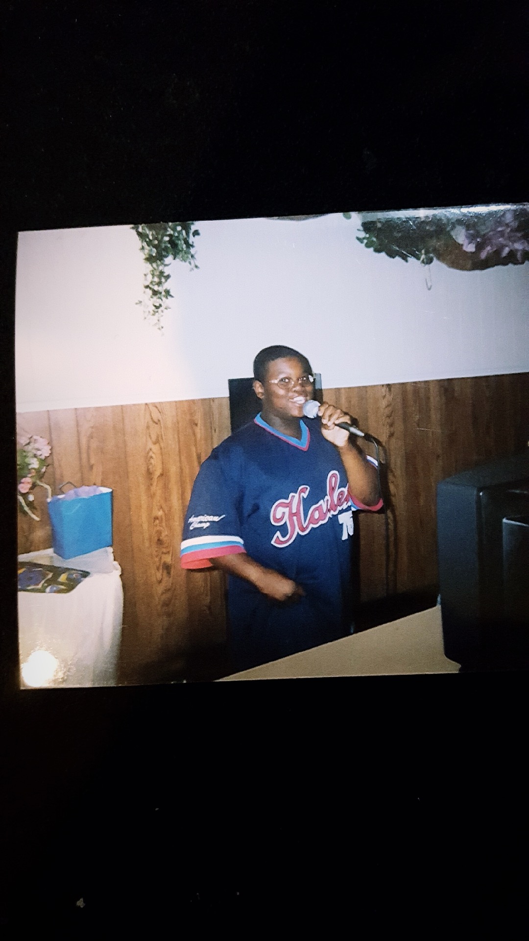 Person wearing a dark blue sports jersey with red and white accents, holding a microphone, standing indoors against a white and wood-paneled wall.