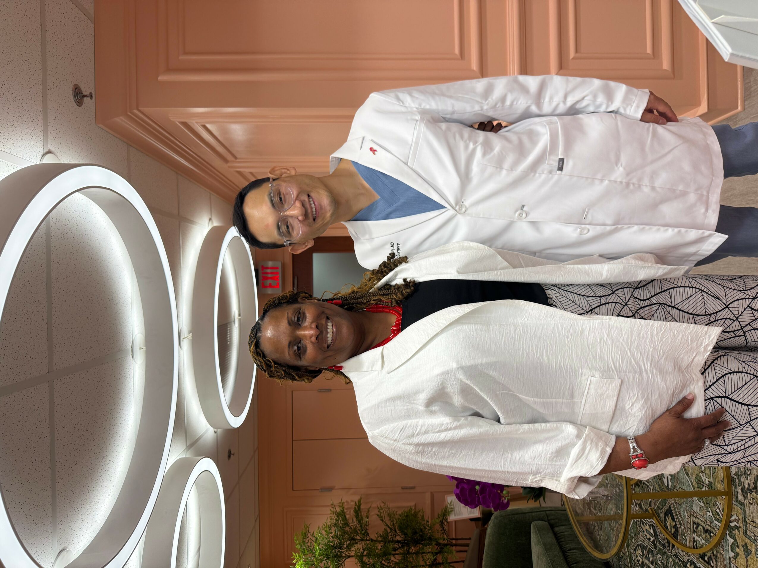 Two women in white coats standing in a medical or dental office with equipment and cabinets behind them.