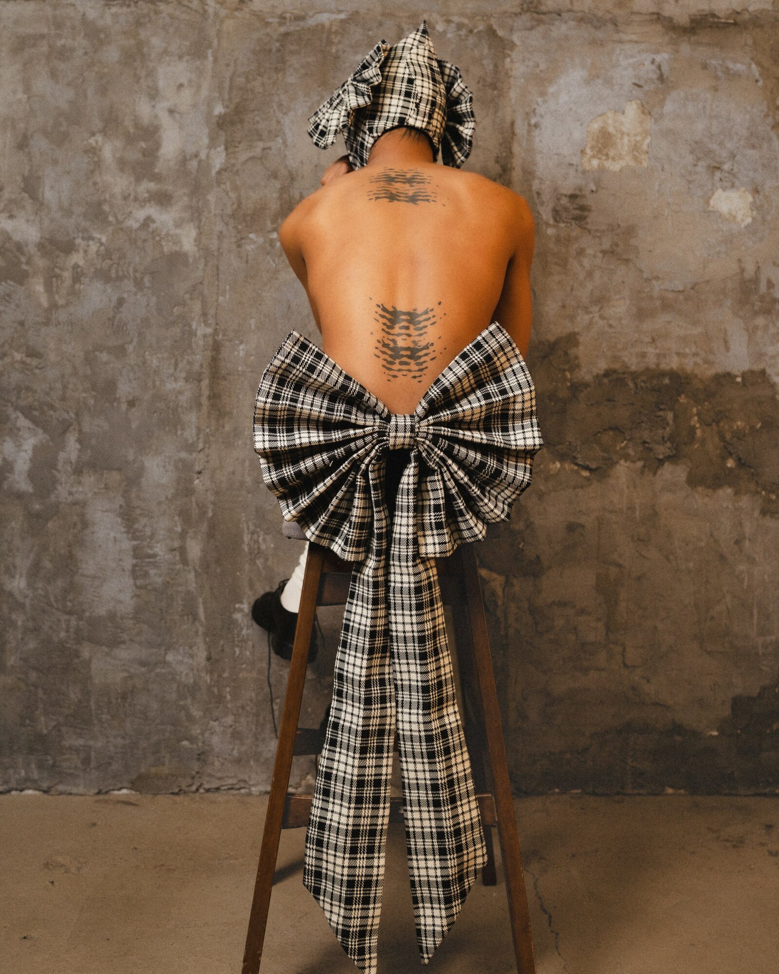 Person with back tattoo sitting on a stool, wearing a large plaid bow and head covering, against a textured wall.