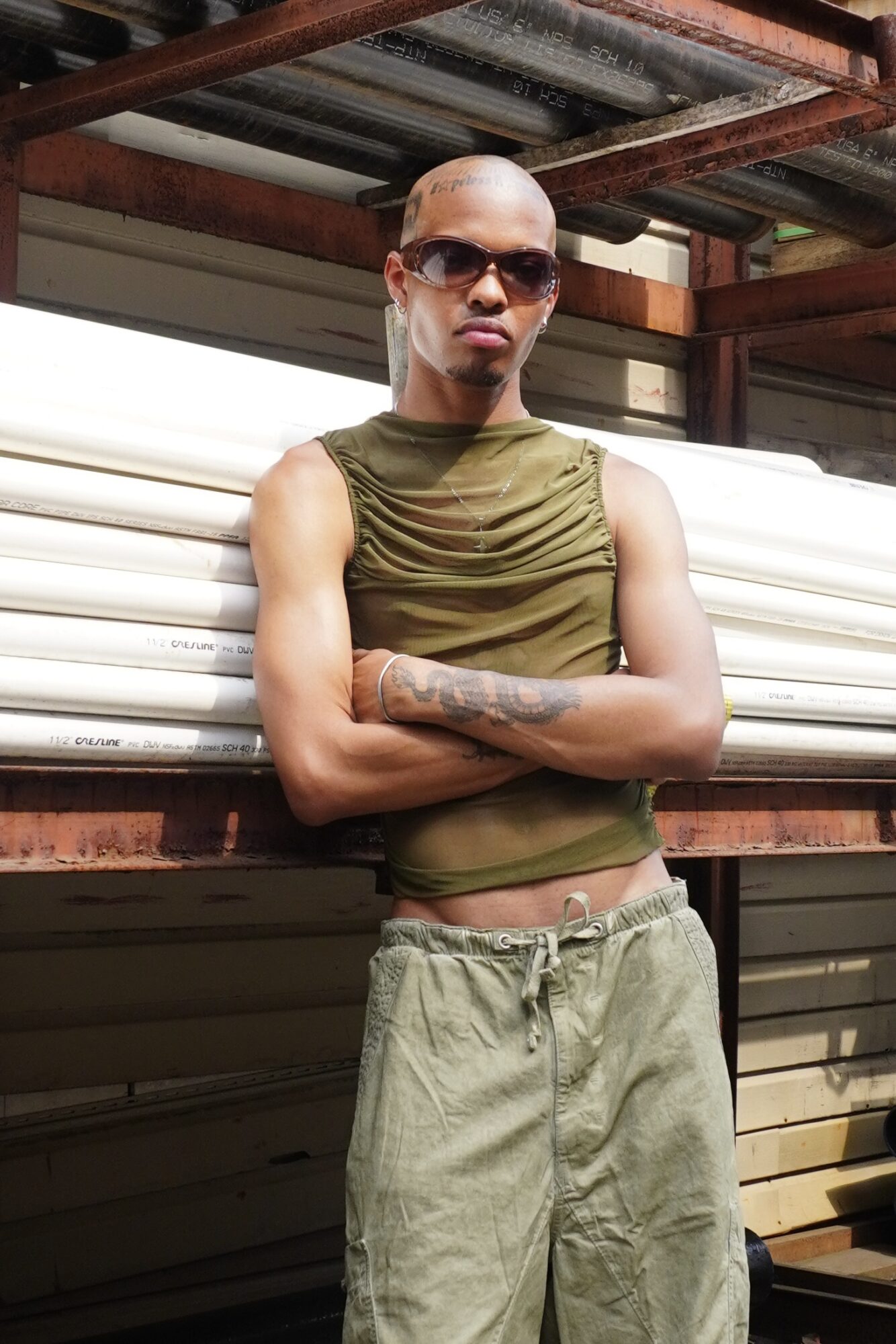 Person with sunglasses, sleeveless shirt, and tattooed arm leaning against a metal structure outdoors.