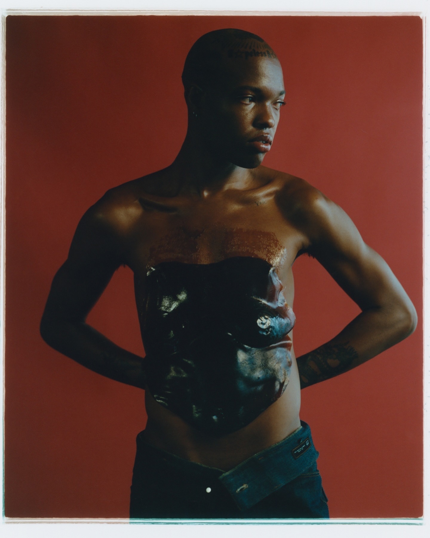 Person with short hair and dark skin wearing a strapless top and jeans, standing against a red background.